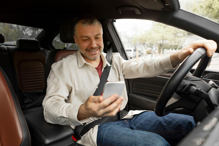 A man looking at his smartphone while driving a car, showing dumb ways people ruin their lives by distracted driving.