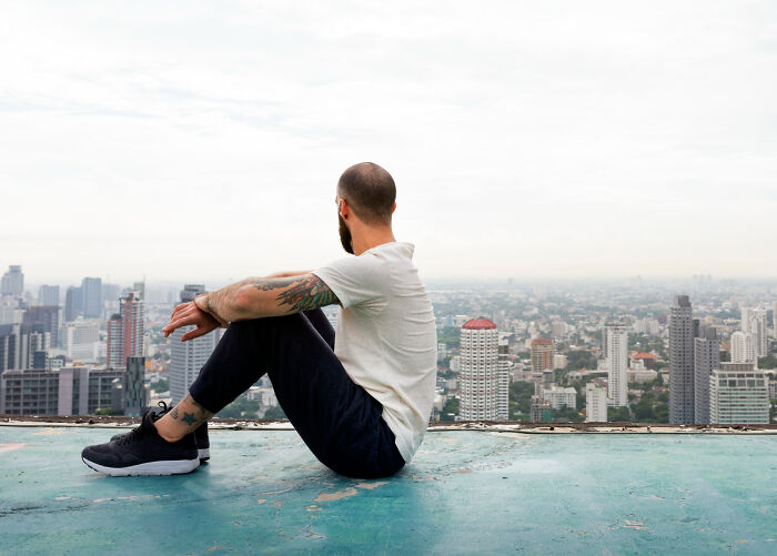 A man with tattoos sitting on a rooftop, overlooking a city, reflecting on an unexpected life story end.