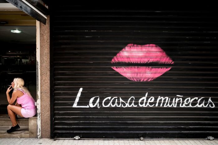 A street photographer captures an odd scene: a woman sitting next to a garage door with pink lips graffiti and text.