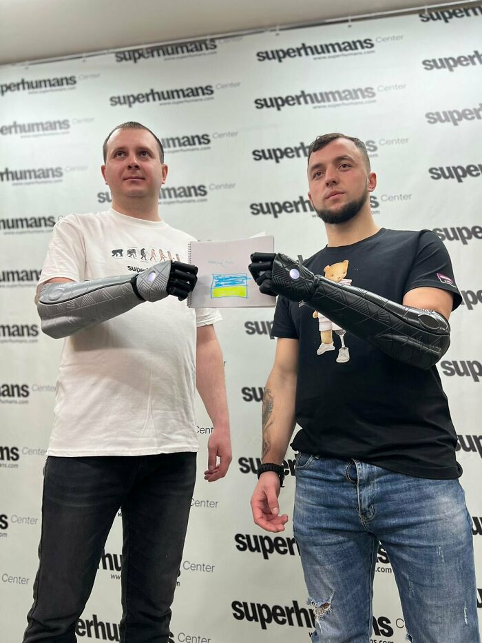 Two men proudly displaying their advanced 3D printers prosthetic arms and a drawing of the Ukrainian flag.