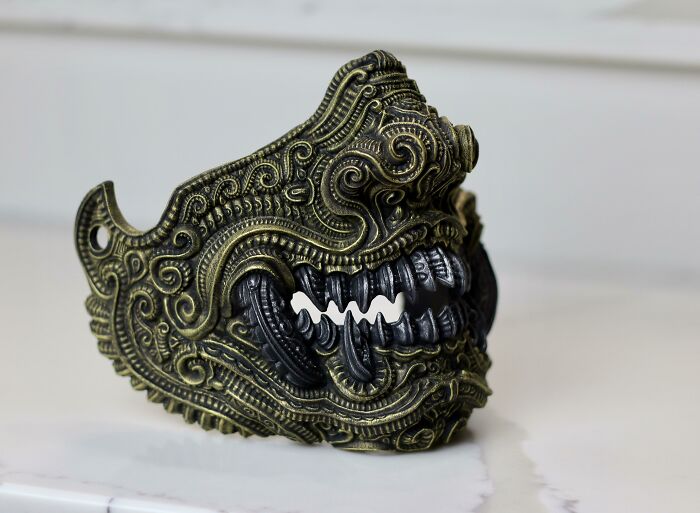 An ornate, gold and black 3D printed mask with intricate details and fierce teeth, a brilliant example of 3D printers.