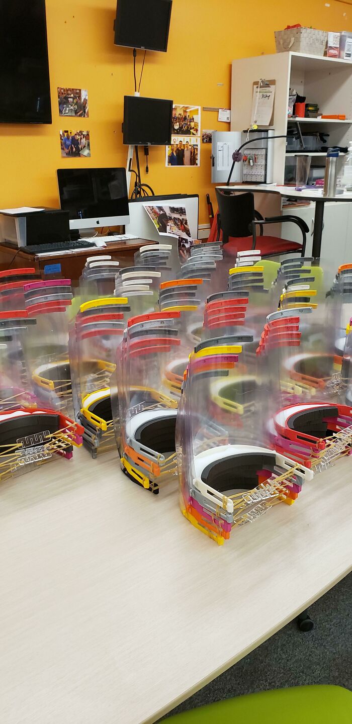 Numerous 3D printed face shields lined up on a table in a classroom or workshop setting.