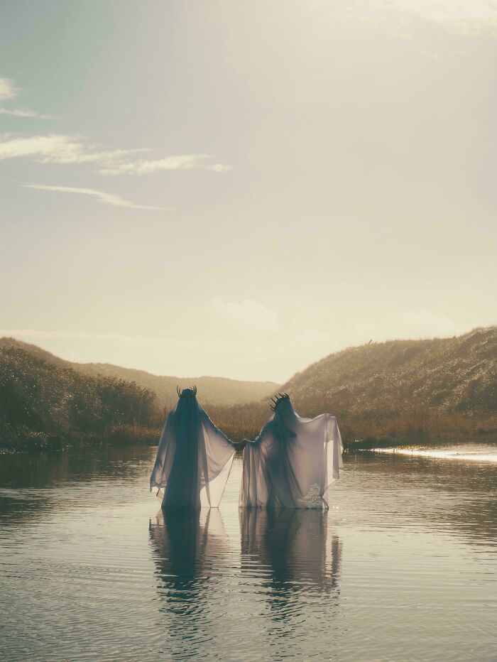 Two ethereal figures in long white robes, brilliant creations from 3D printers, stand in water at sunset.