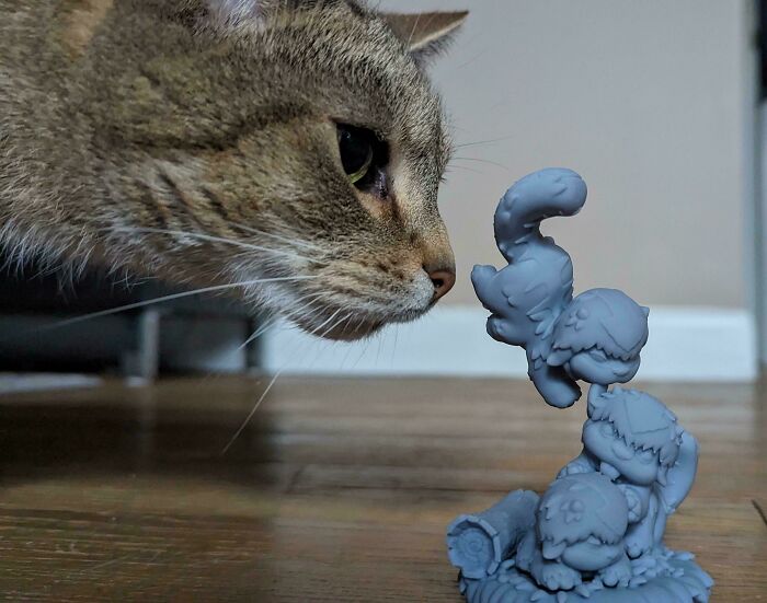 A curious cat observing a small, detailed gray figurine, brilliant stuff created with 3D printers, on a wooden floor.