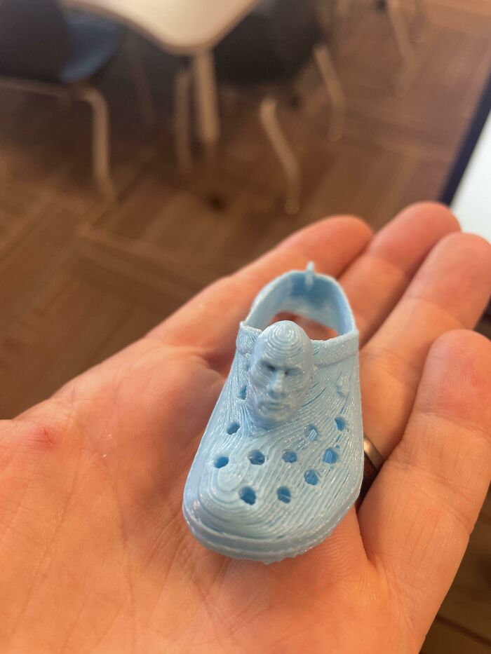A hand holding a tiny, light blue 3D printed Croc shoe with a man's face on the front, brilliant stuff.