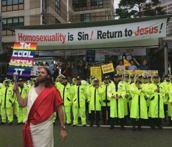 A person dressed as Jesus holding a rainbow flag, standing in front of a protest banner that reads 'Homosexuality is Sin! Return to Jesus!'