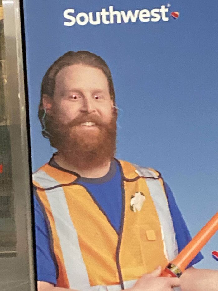 A man with a beard wearing a safety vest smiling in front of a Southwest Airlines ad at the airport, a weird thing.