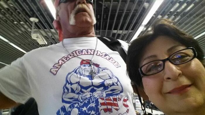 A man resembling Hulk Hogan wearing an American Made shirt poses for a selfie at an airport, a hilarious thing spotted at airports.