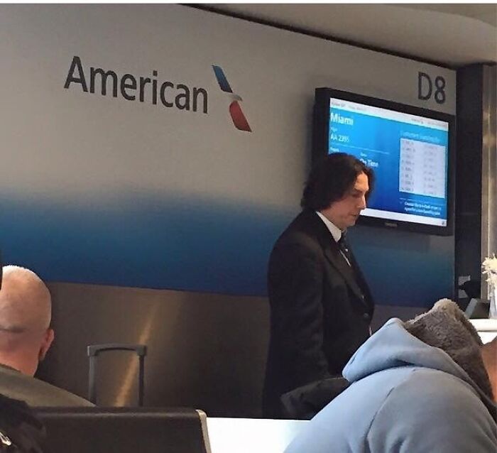 A man with long dark hair in a suit standing at an American Airlines counter, one of the weird things spotted at airports.