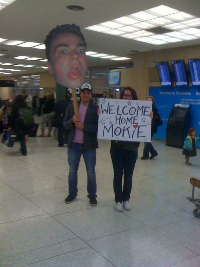 Two people at the airport holding a large head cutout and a 'Welcome Home Mokie' sign. Hilarious and weird things spotted at airports.
