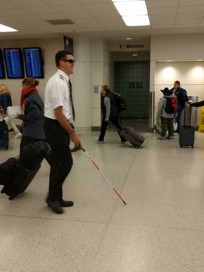 A man in a pilot uniform with dark sunglasses and a white cane walking through the airport. Hilarious and weird things spotted at airports.