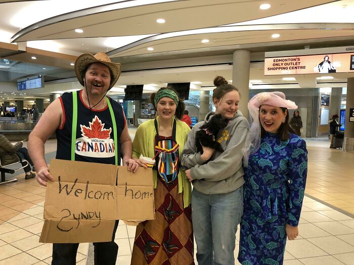 Four people in a colorful welcome group, including a man in a Canadian shirt holding a 'Welcome home' sign, at the airport. Hilarious and weird things spotted at airports.