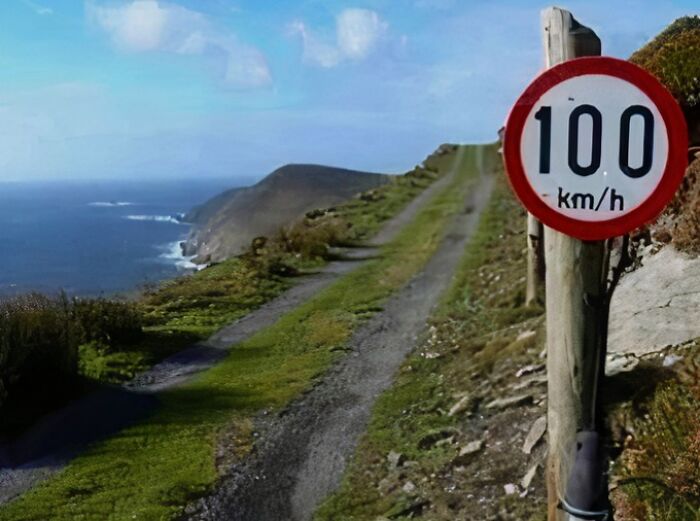 A funny sign with a 100 km/h speed limit on a very narrow, unpaved road alongside a cliff overlooking the ocean.