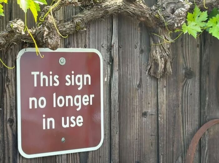 A silly funny sign that reads This sign no longer in use, hanging on a wooden fence with green vines.