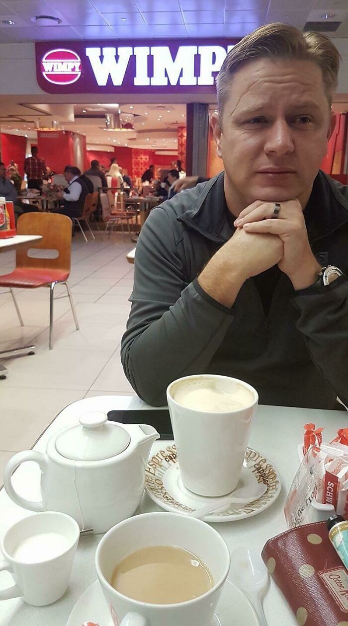 A man with blond hair resting his chin on his hands, sitting at a table with coffee, a weird thing spotted at airports.