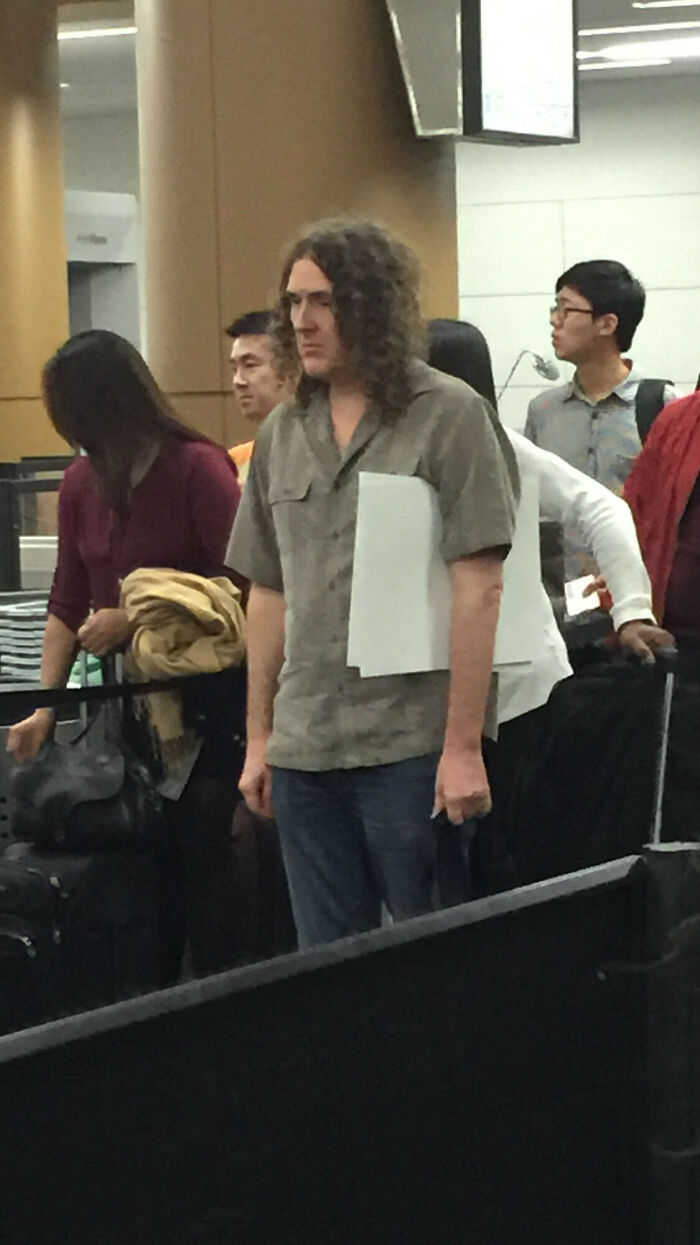 A hilarious thing spotted at airports: a man with long curly hair and a grey shirt holding a piece of paper.