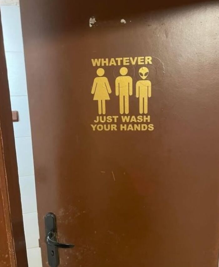 A silly funny sign on a brown door shows stick figures for male, female, and alien with text Whatever, Just Wash Your Hands.