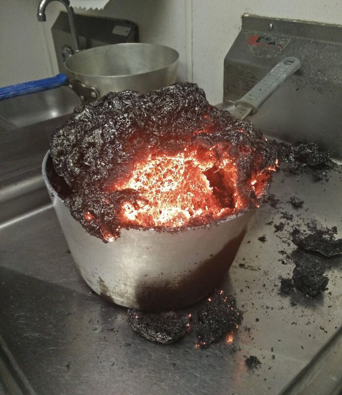 A dramatic kitchen fail with a pot of burnt food glowing red hot, making a disastrous mess in a metal sink.
