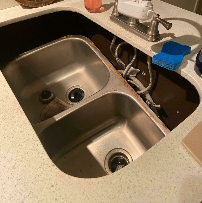 A double kitchen sink has fallen out of the countertop, showing severe kitchen fails.