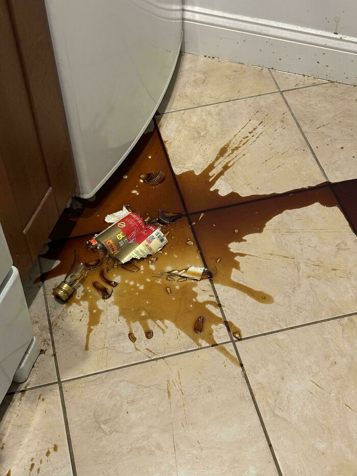 A broken bottle and spilled liquid on a tiled kitchen floor, creating a messy kitchen fail.