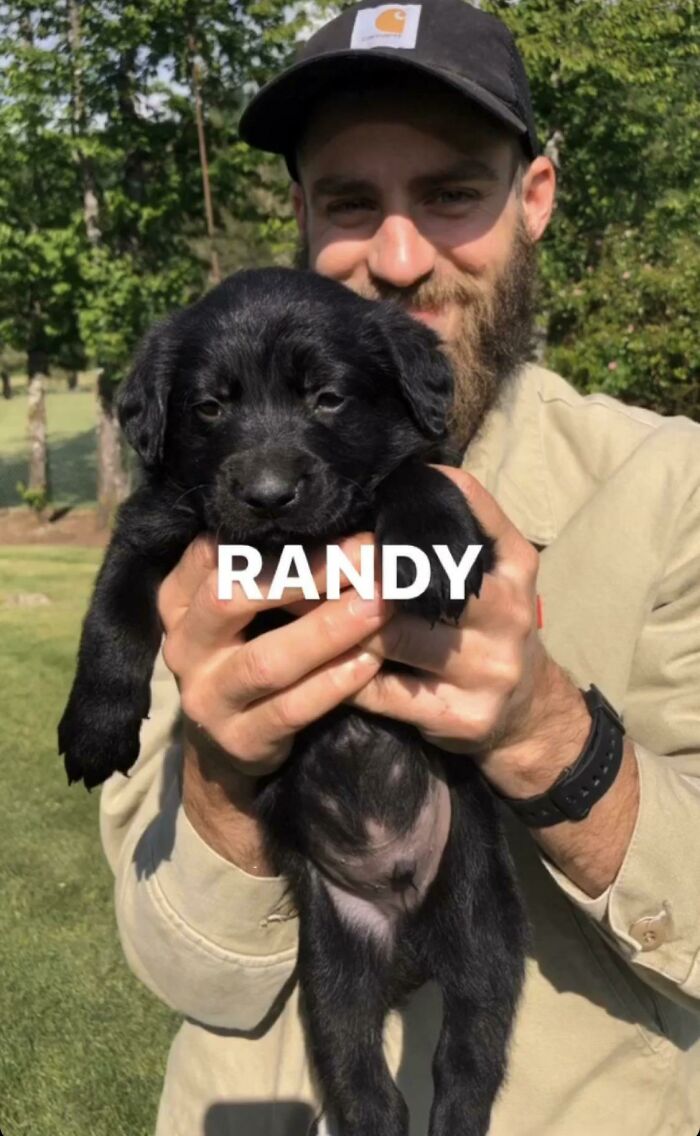 A smiling man in a cap and beard holding a small black puppy named Randy, a wholesome animal photo.