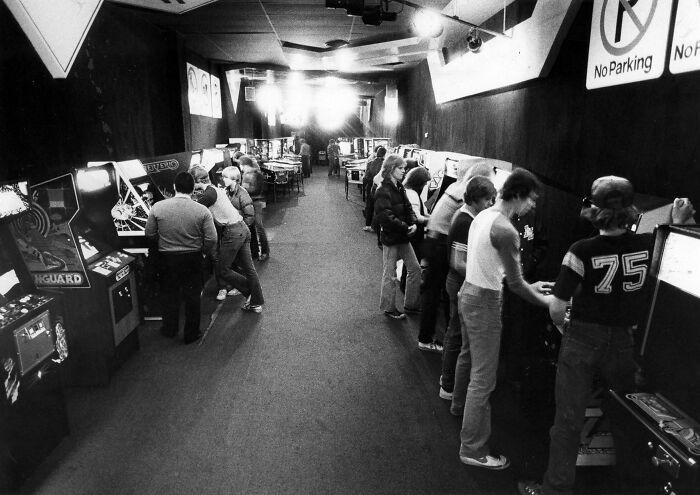 A black and white photo of an arcade with people playing various games, a scene of retro tech from the 80s.