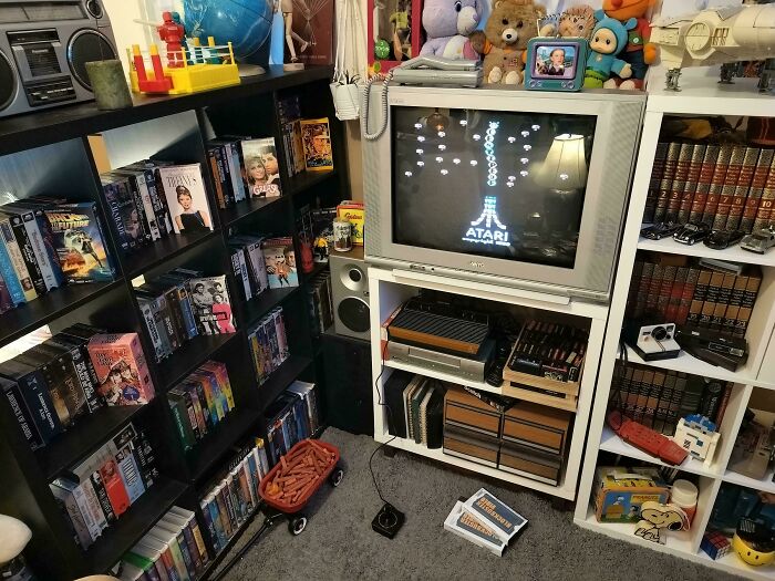 A room filled with 80s retro tech, including an Atari console connected to a CRT TV, VHS tapes, and bookshelves.