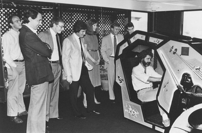 A group of men observing a person playing a Star Wars arcade game, highlighting the popularity of 80s retro tech.