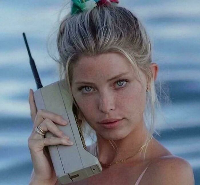 A woman with blonde hair holds a large, grey mobile phone to her ear, embodying the style of 80s retro tech communications.