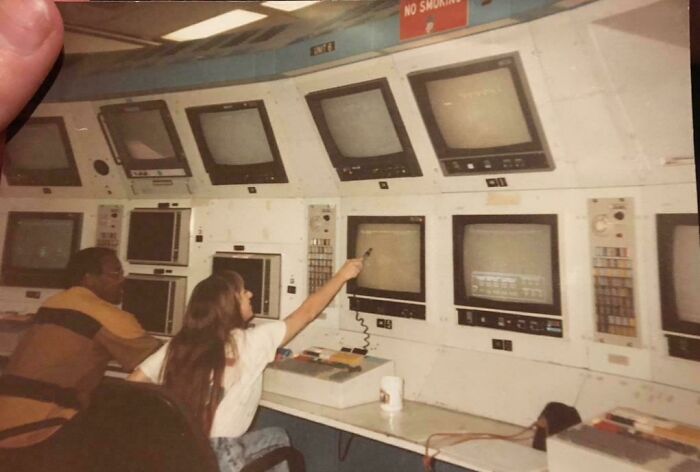 Two individuals in an '80s control room surrounded by retro tech monitors.