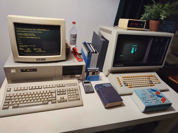Two retro tech computer setups on a desk with keyboards, monitors, and floppy disks, from the 80s.