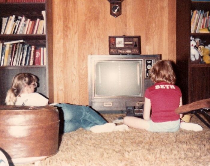 Two girls on the floor in front of a retro tech TV, enjoying 80s entertainment in a living room.