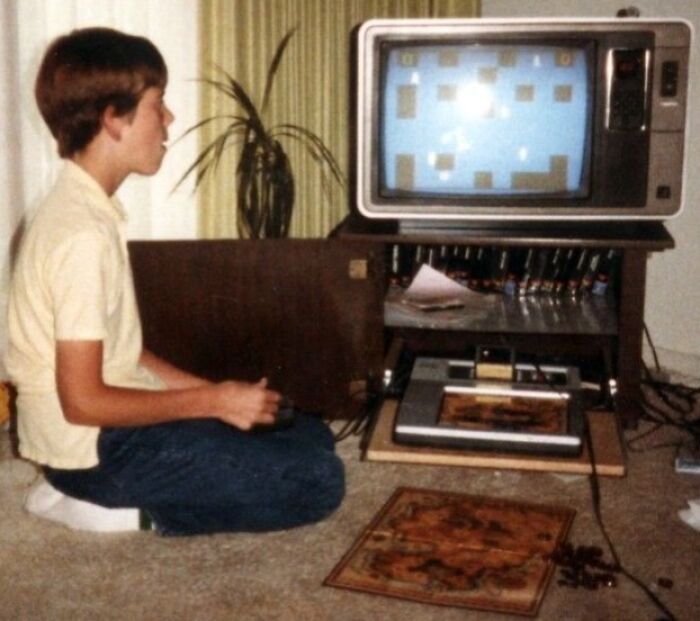 A young boy intently plays a video game on a classic TV with '80s retro tech, sitting on the floor.