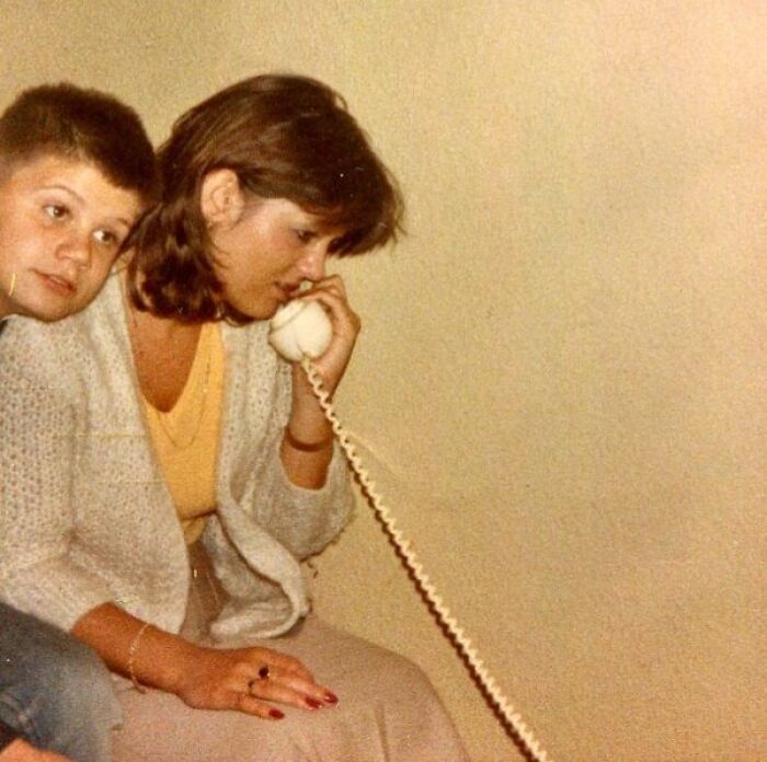 A woman talks on a retro tech phone with a long coiled cord, while a child sits beside her, capturing the 80s weirdness.