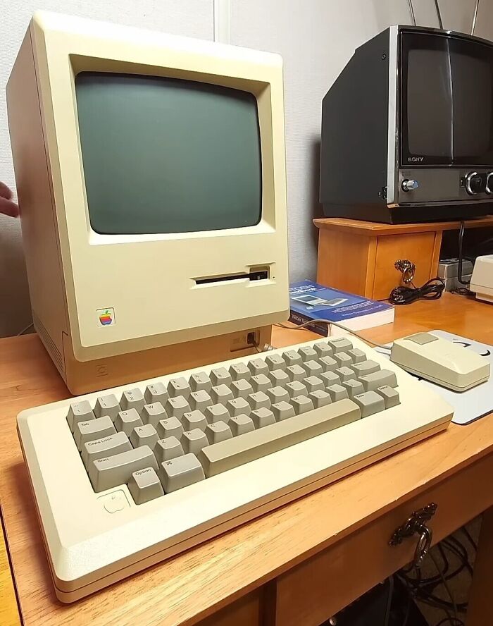 A classic Macintosh computer setup on a wooden desk, showcasing 80s retro tech with a CRT monitor, keyboard, and mouse.