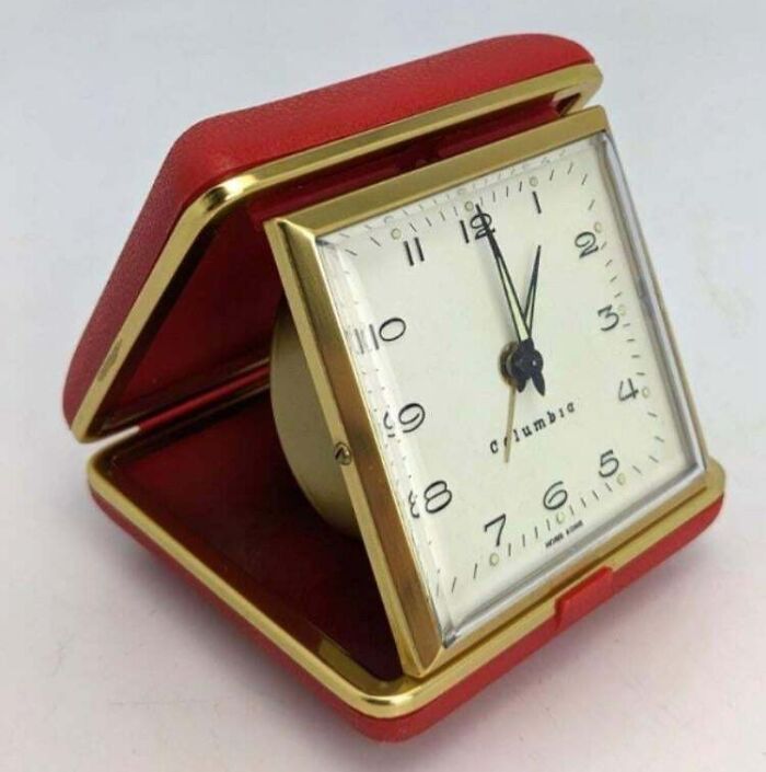 A small, red and gold travel alarm clock, an iconic 80s retro tech item, with a cream-colored face displaying the time.