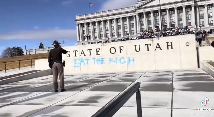 A police officer stands in front of a government building with 'Eat The Rich' spray-painted on the wall, showing a protest moment.