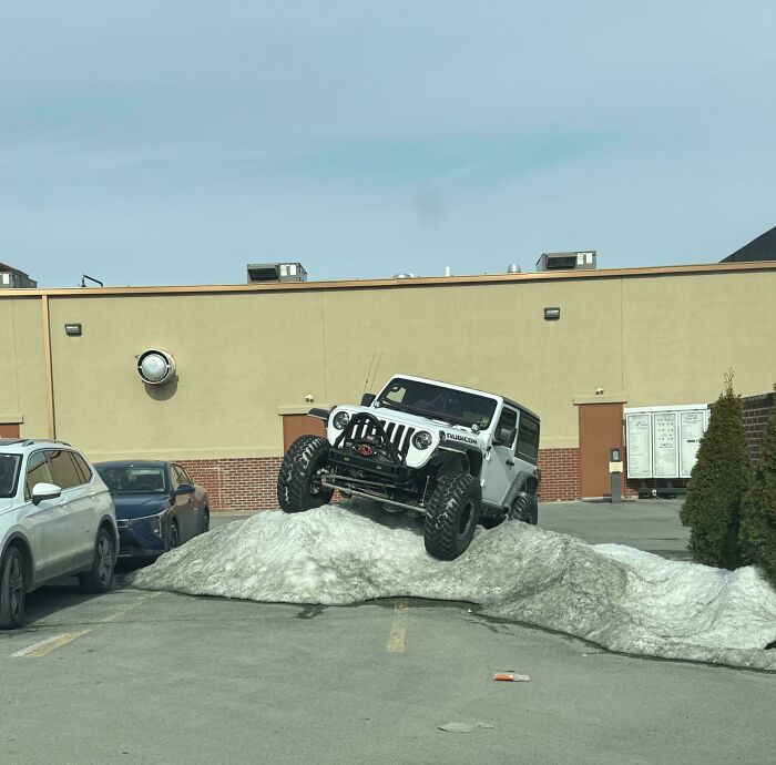 A white Jeep Rubicon on a pile of snow in a parking lot, showing Americans doing something stupid.