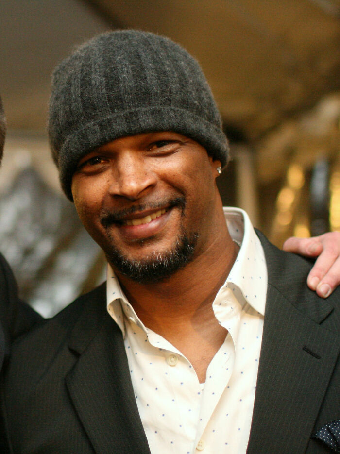 Damon Wayans smiling, wearing a dark beanie, a white polka-dot shirt, and a dark suit jacket, highlighting his career.