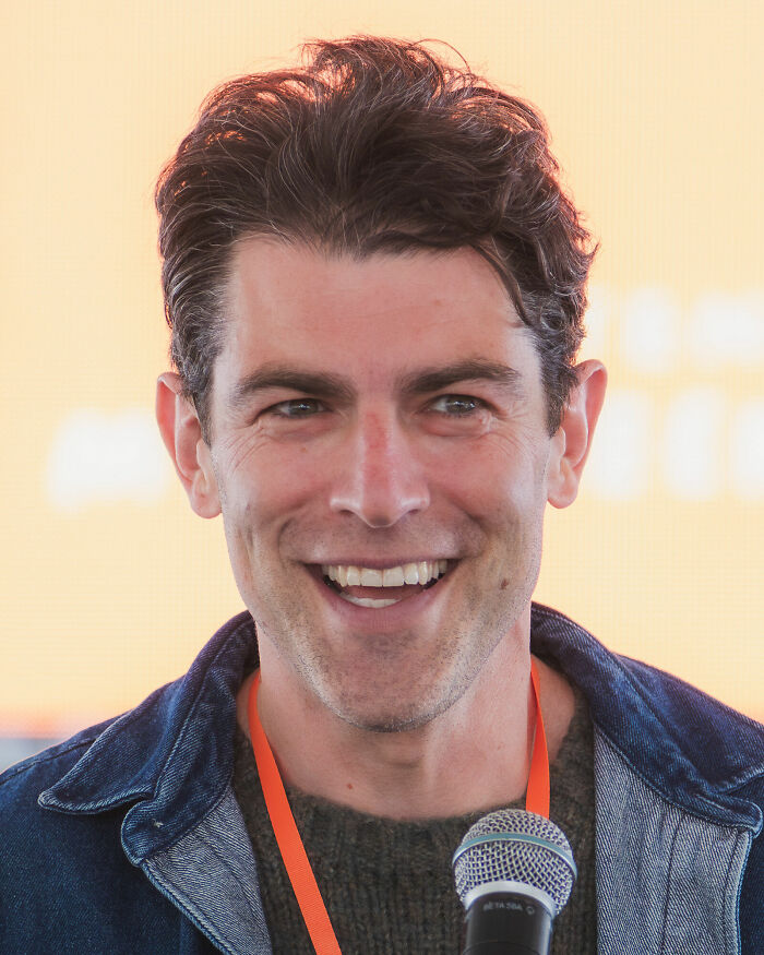 Max Greenfield smiling into a microphone, showcasing his bio and career highlights.