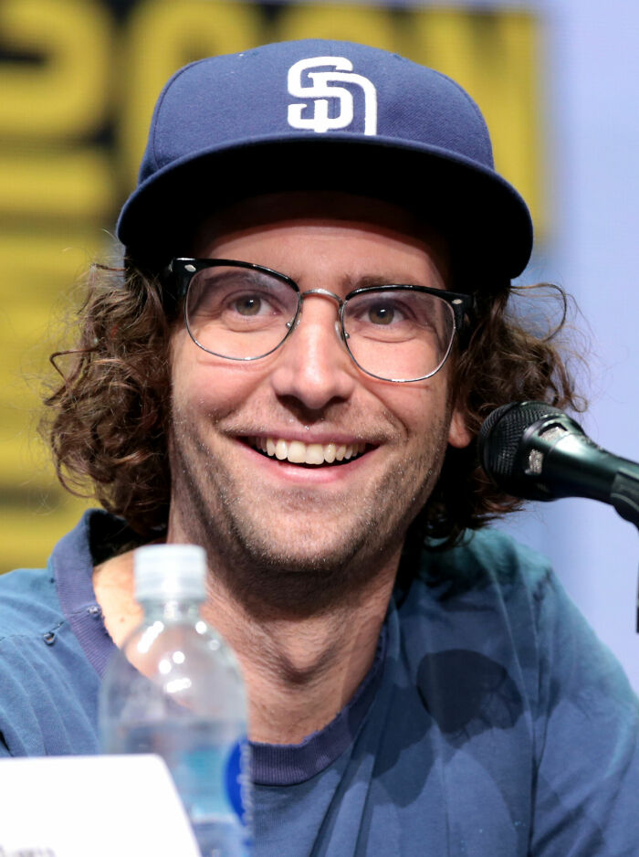 A close-up of Kyle Mooney smiling, wearing glasses and a blue baseball cap, highlighting his career highlights.