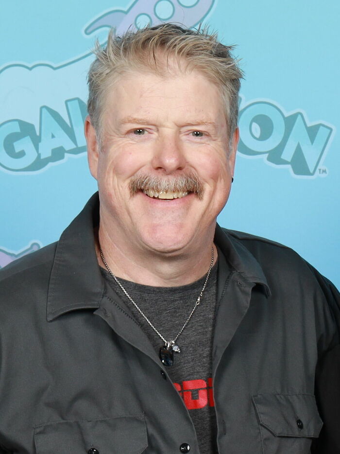 John DiMaggio smiling, showcasing his career highlights and bio. He has a mustache and wears a gray shirt with a necklace.