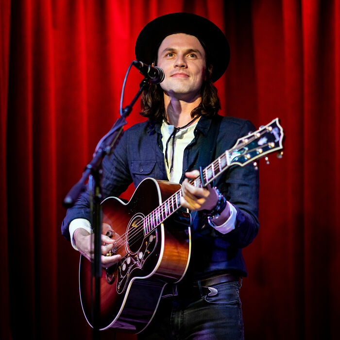 James Bay performing live, showcasing his career highlights with an acoustic guitar and a black hat on a red stage.