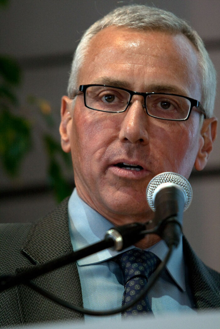 Drew Pinsky in glasses speaking into a microphone, highlighting his bio and career achievements.