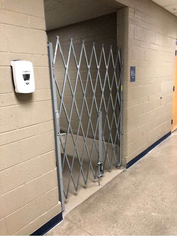 A locked collapsible gate blocks a public restroom, indicating the horrid and sad world with restricted access.