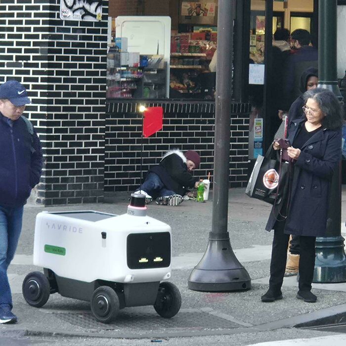 A delivery robot passes by a person experiencing homelessness, showing a horrid world disparity.