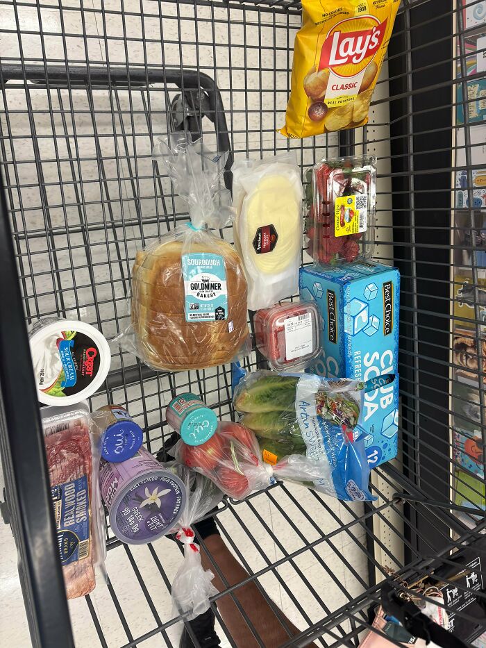 A shopping cart filled with groceries, including bread, produce, and packaged foods, showing the cost of living.