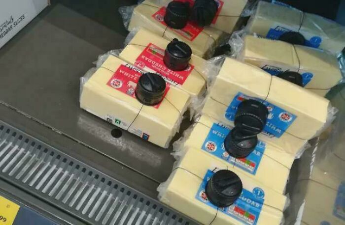 A supermarket aisle displays cheese blocks fitted with security devices, reflecting the horrid world we're living in.