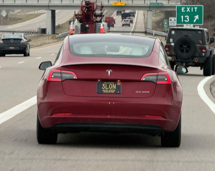 A red Tesla with a vanity license plate 3L0N, on a highway, part of the horrid and sad world.