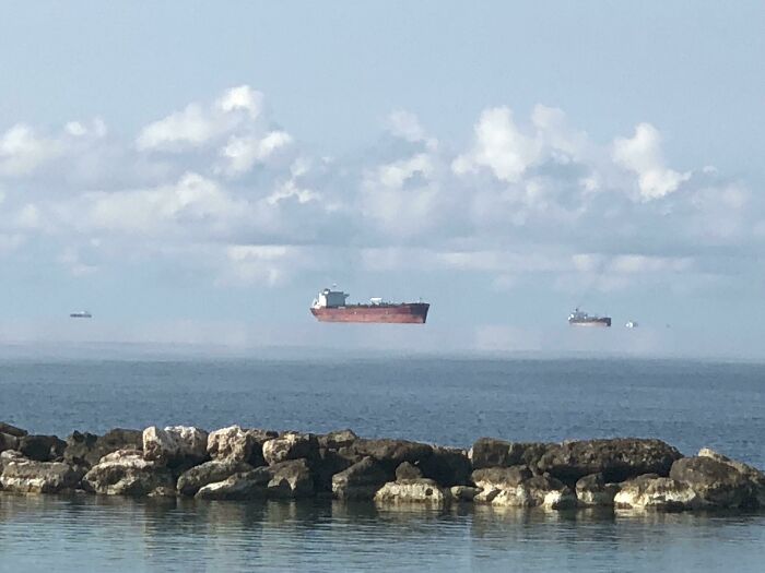 Several large ships appear to be floating above the water's surface on a cloudy day, a baffling image of physical illusion.